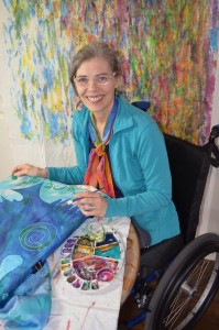 image shows a smiling woman in colourful clothes painting in a wheelchair